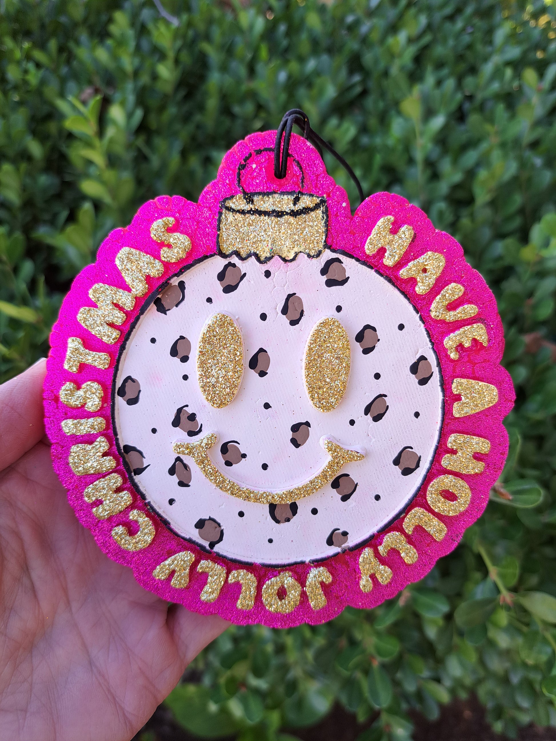 leopard print Christmas bulb freshie that says holly jolly. Held in hand in front of shrub