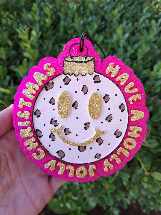 leopard print Christmas bulb freshie that says holly jolly. Held in hand in front of shrub