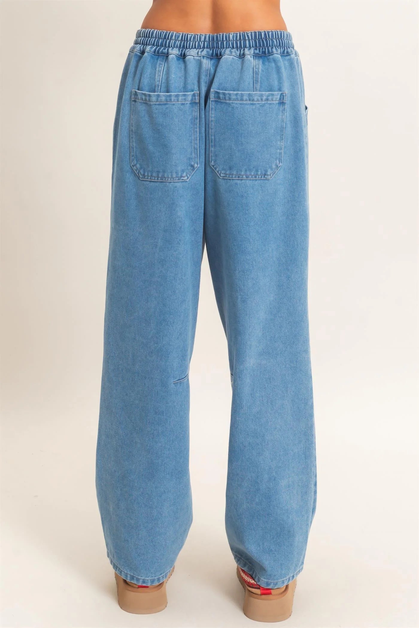 Rear view of a person wearing light blue wide-leg denim pants with an elastic waistband and two back patch pockets, shown against a plain light beige studio background.