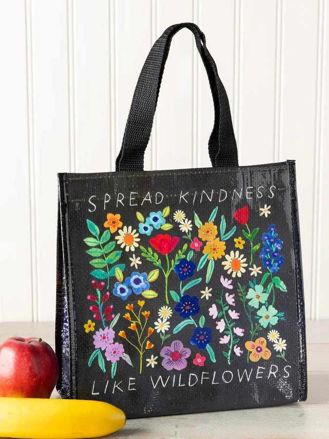 A black insulated tote bag with colorful wildflower illustrations and the text 'SPREAD KINDNESS LIKE WILDFLOWERS' on the front, shown standing upright with an apple and banana at the base against a white paneled wall.