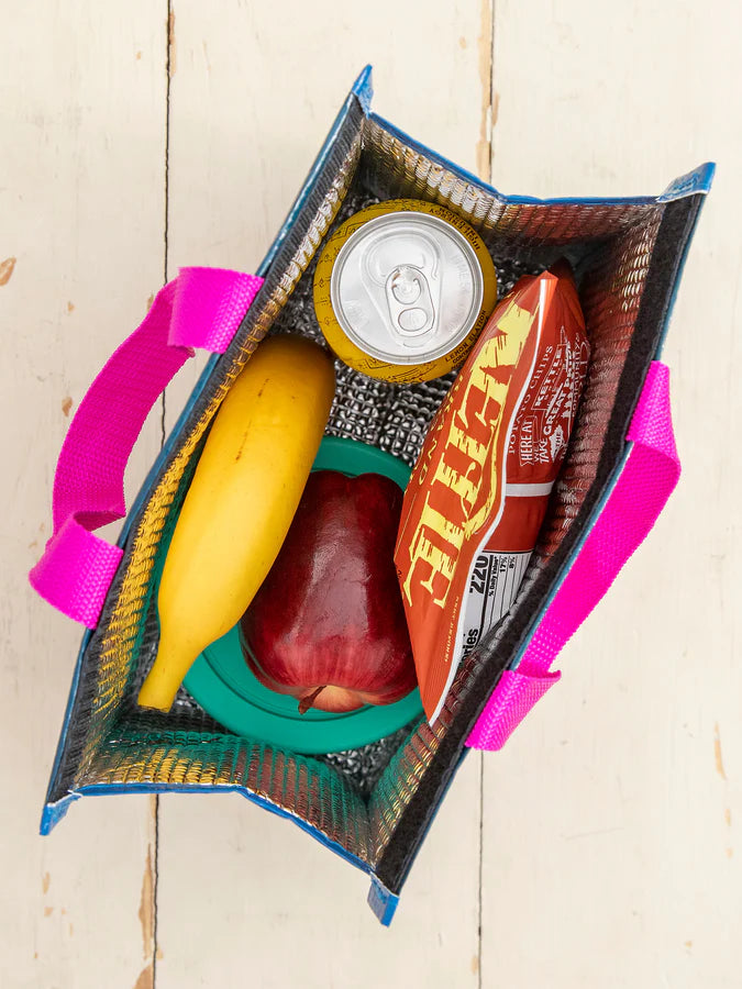 A reusable insulated lunch bag with bright pink handles, shown from above on a light wood surface. Inside are a banana, a red apple, a green container, a can, and a red snack package.