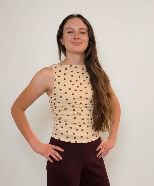 woman wearing an ivory and brown polka dot mesh top with brown pants on a neutral background