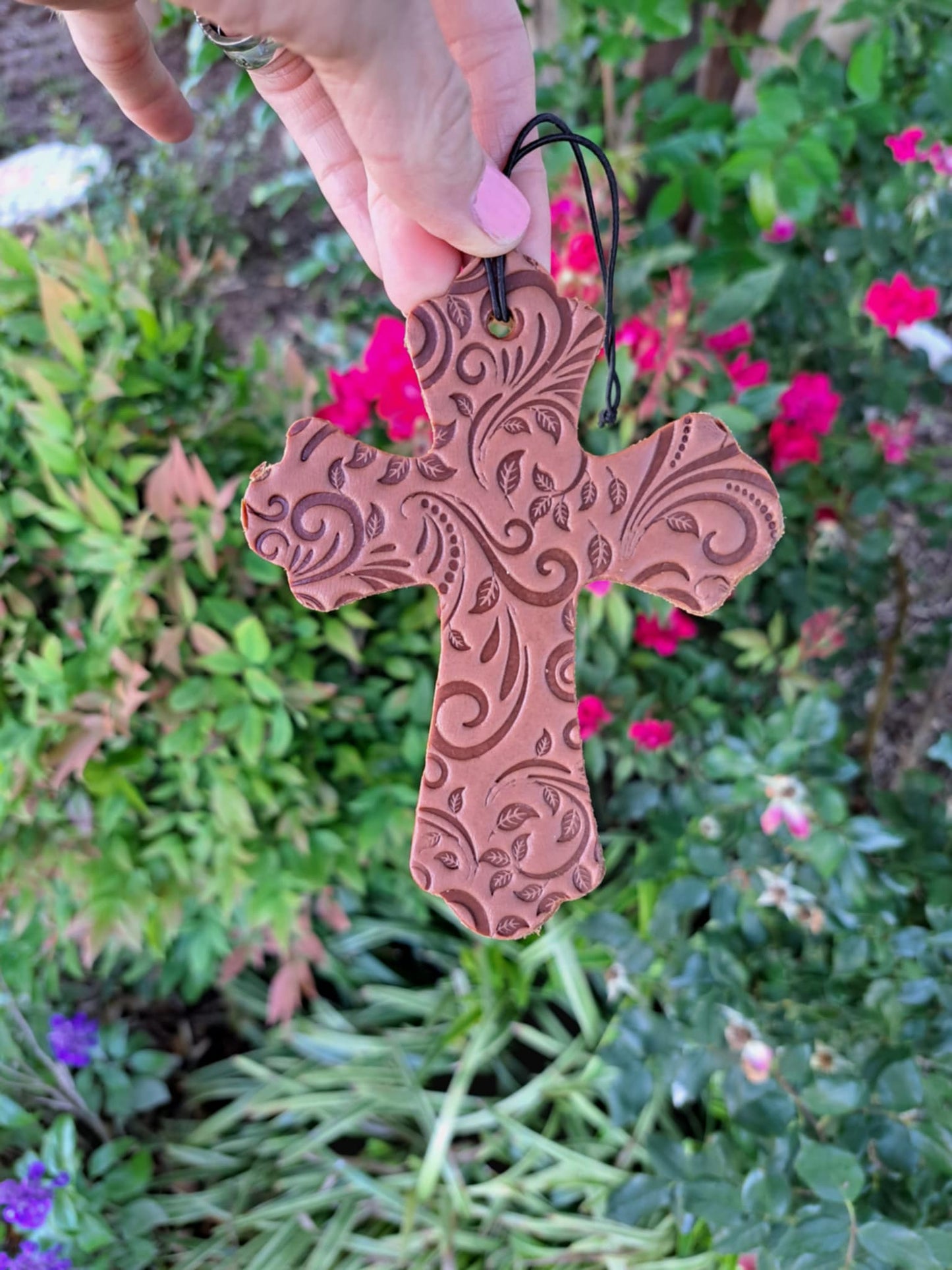 A tooled leather tooled cross freshie hanging on an elastic string against a background of green plants and pink flowers.