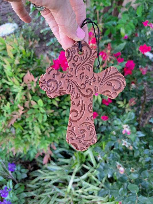 A tooled leather tooled cross freshie hanging on an elastic string against a background of green plants and pink flowers.