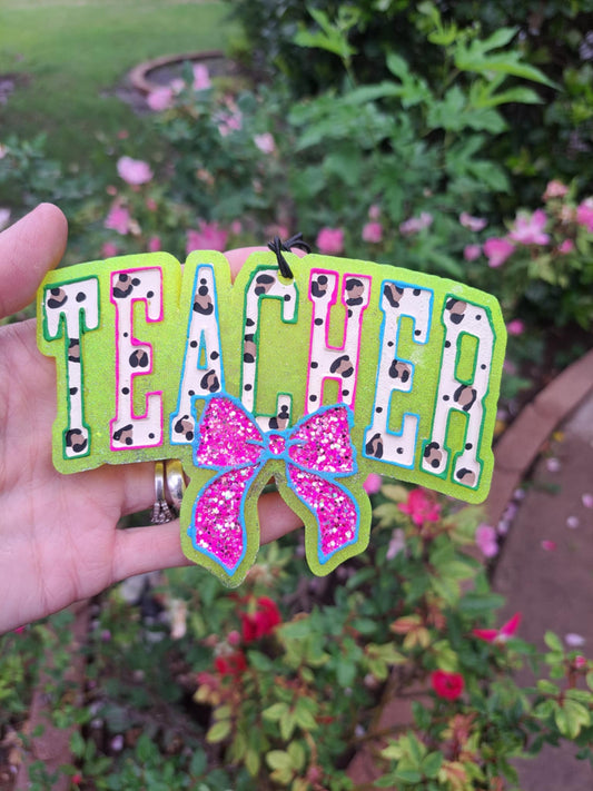 Handmade and painted hanging air freshener with the word 'TEACHER' in a colorful outline, attached to a pink glitter bow and a black elastic string, set against a floral background.