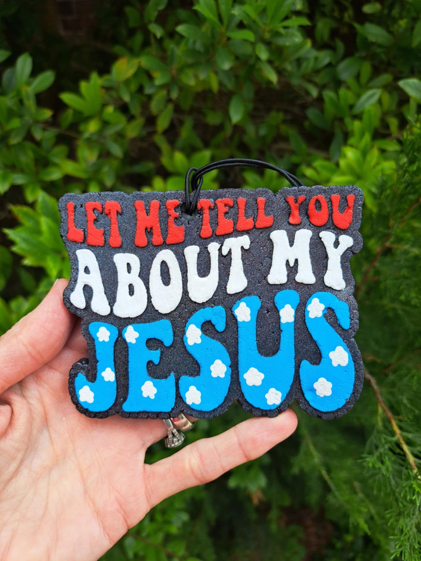 Red, white, and blue freshie that says, 'Let me tell you about my Jesus' message held against a green leafy background