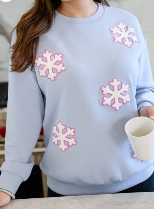 light blue pullover on a woman holding a coffee cup. Top has white snowflake patches with pink sequin trim.