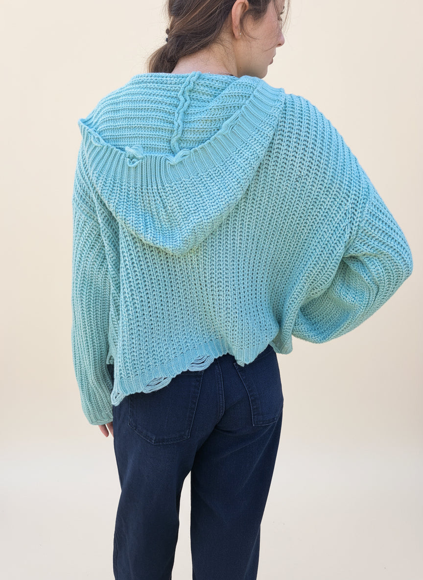 person wearing a mint distressed sweater jacket with dark pants on a cream background