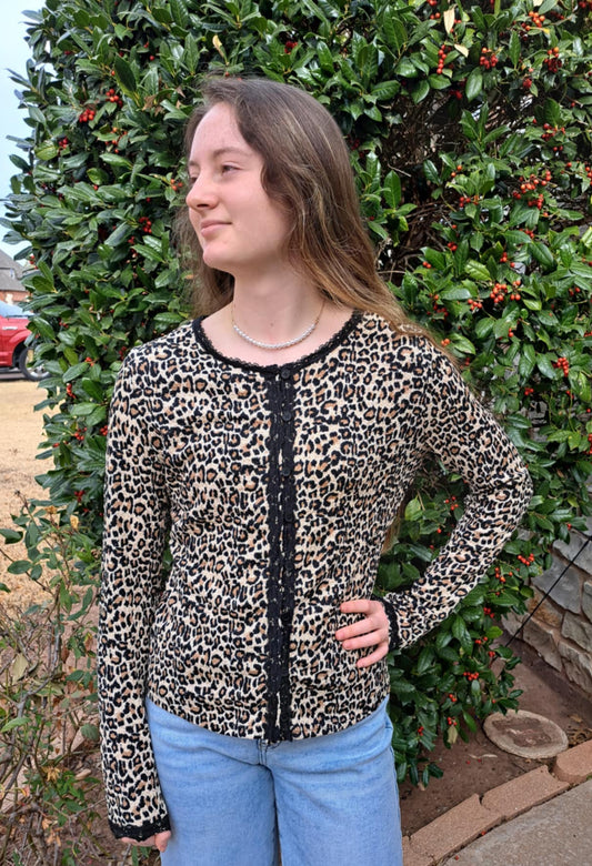 Person wearing a leopard print top and blue jeans standing in front of a stone wall and greenery.