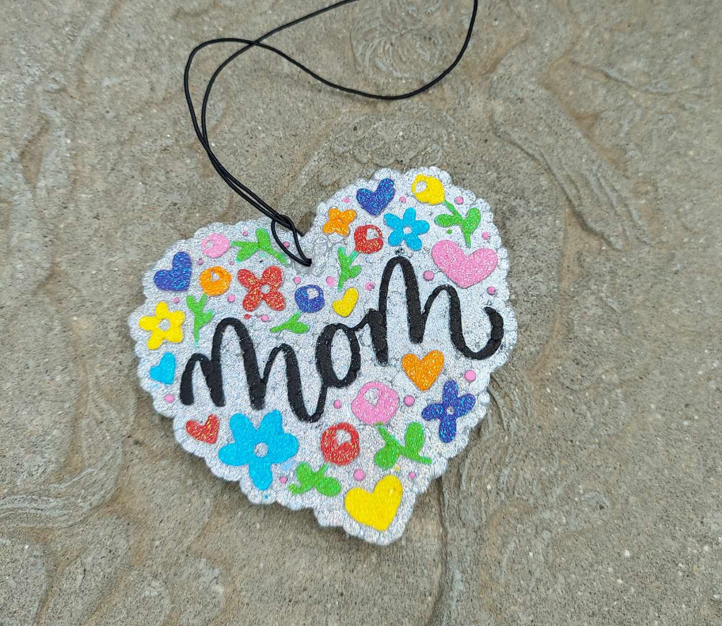 colorful heart air freshener with flowers says MOM. layin on cement park bench