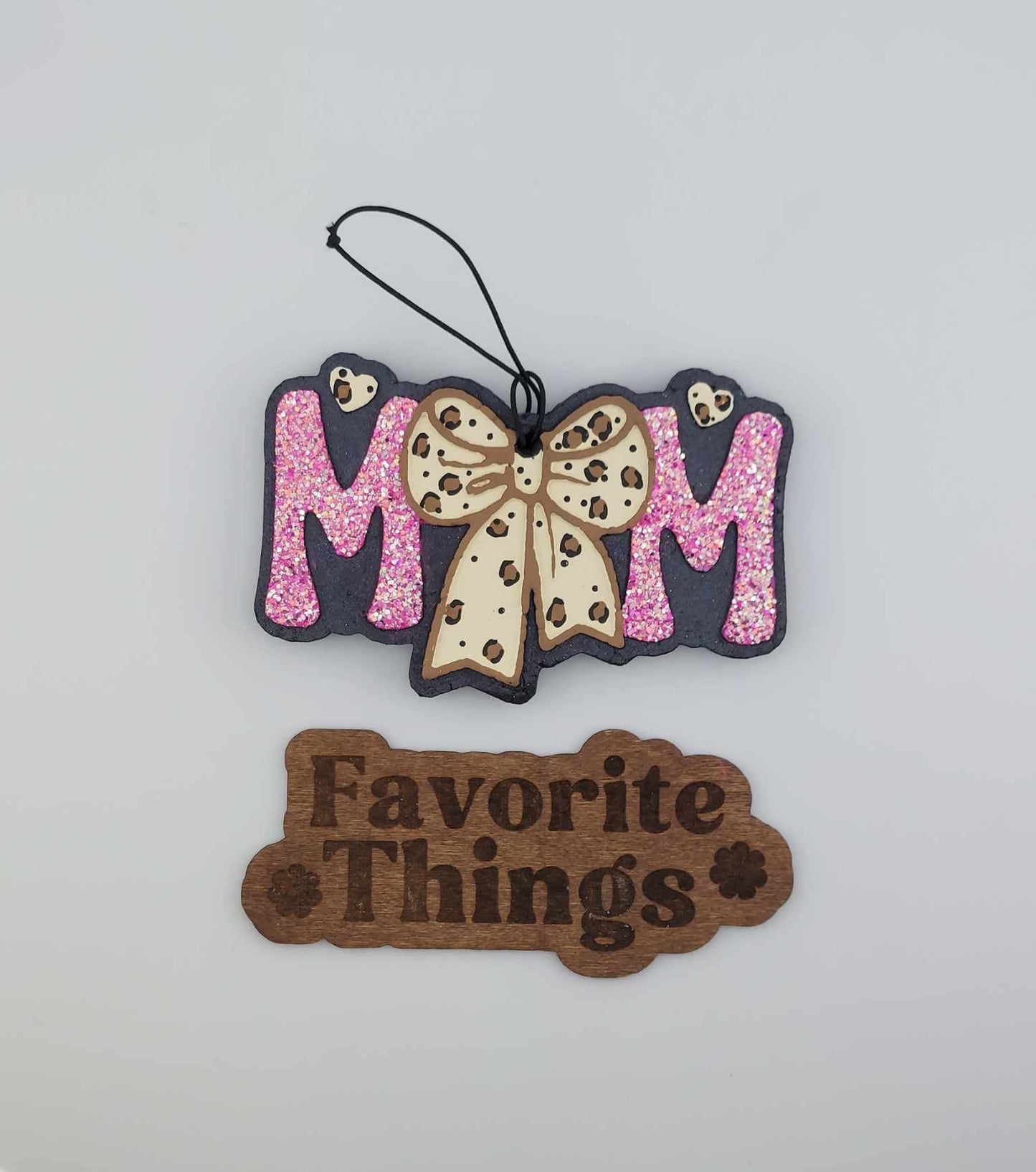 A handmade hanging air freshener with pink glitter and a leopard bow design, labeled 'MOM' at the top and 'Favorite Things' sign at the bottom.