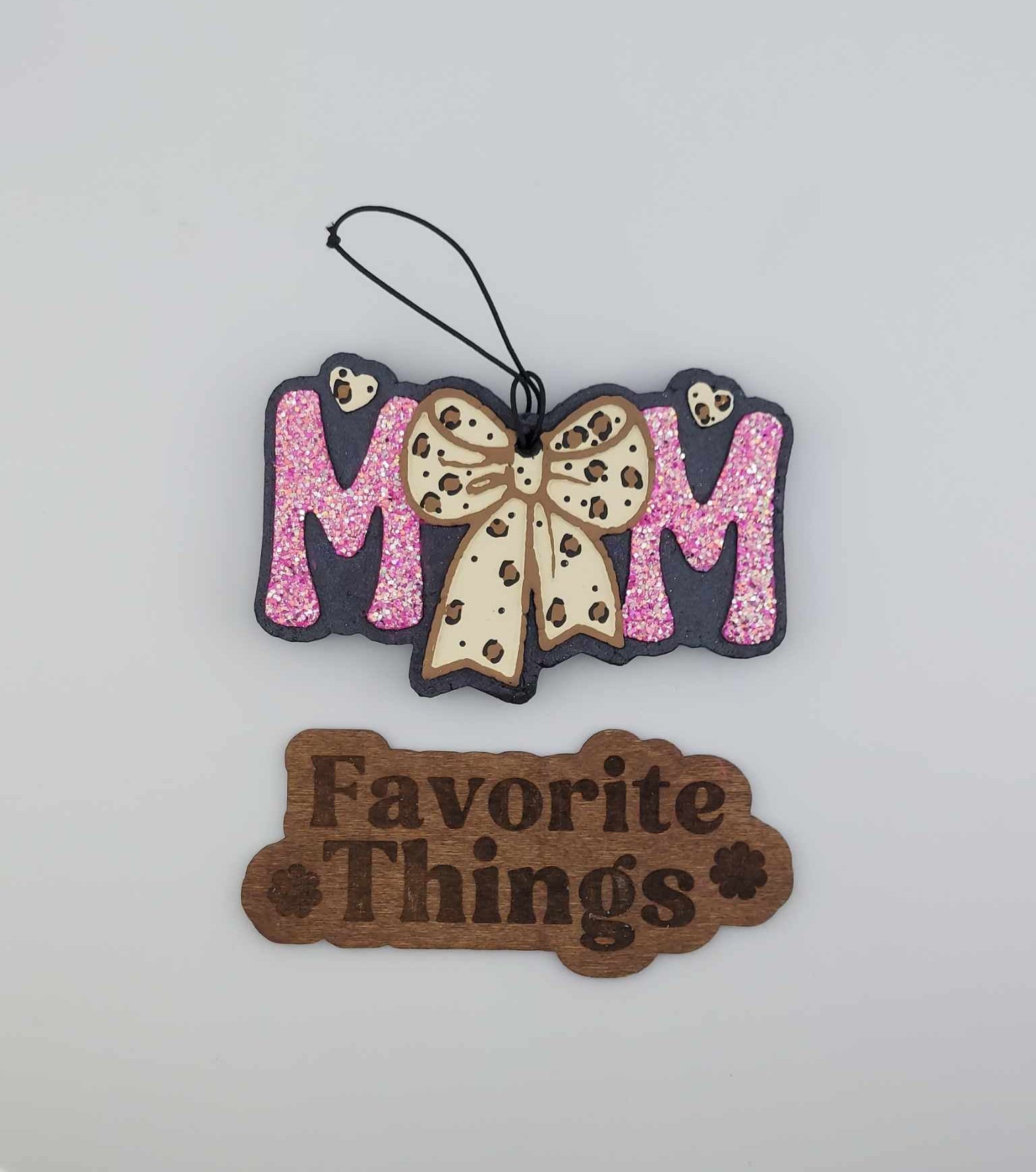 A handmade hanging air freshener with pink glitter and a leopard bow design, labeled 'MOM' at the top and 'Favorite Things' sign at the bottom.