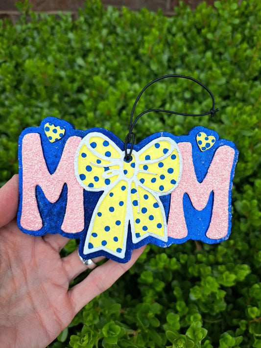 Freshie with 'MOM' and a yellow polka dot bow held against a green leafy background
