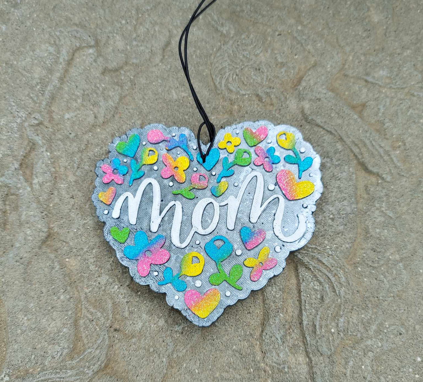A heart-shaped air freshener with 'mom' written in the center, decorated with multicolored ombre flowers on a black elastic string.