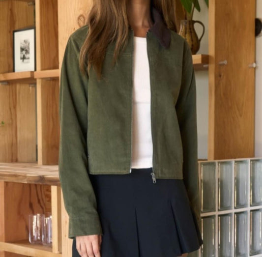 Person wearing a green jacket over a white shirt and black skirt in a room with wooden shelves.