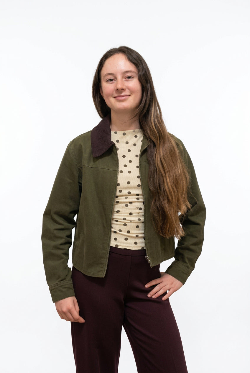 A woman stands in a hallway wearing a dark olive green bomber jacket over a cream top with black polka dots, paired with dark burgundy wide-leg pants.