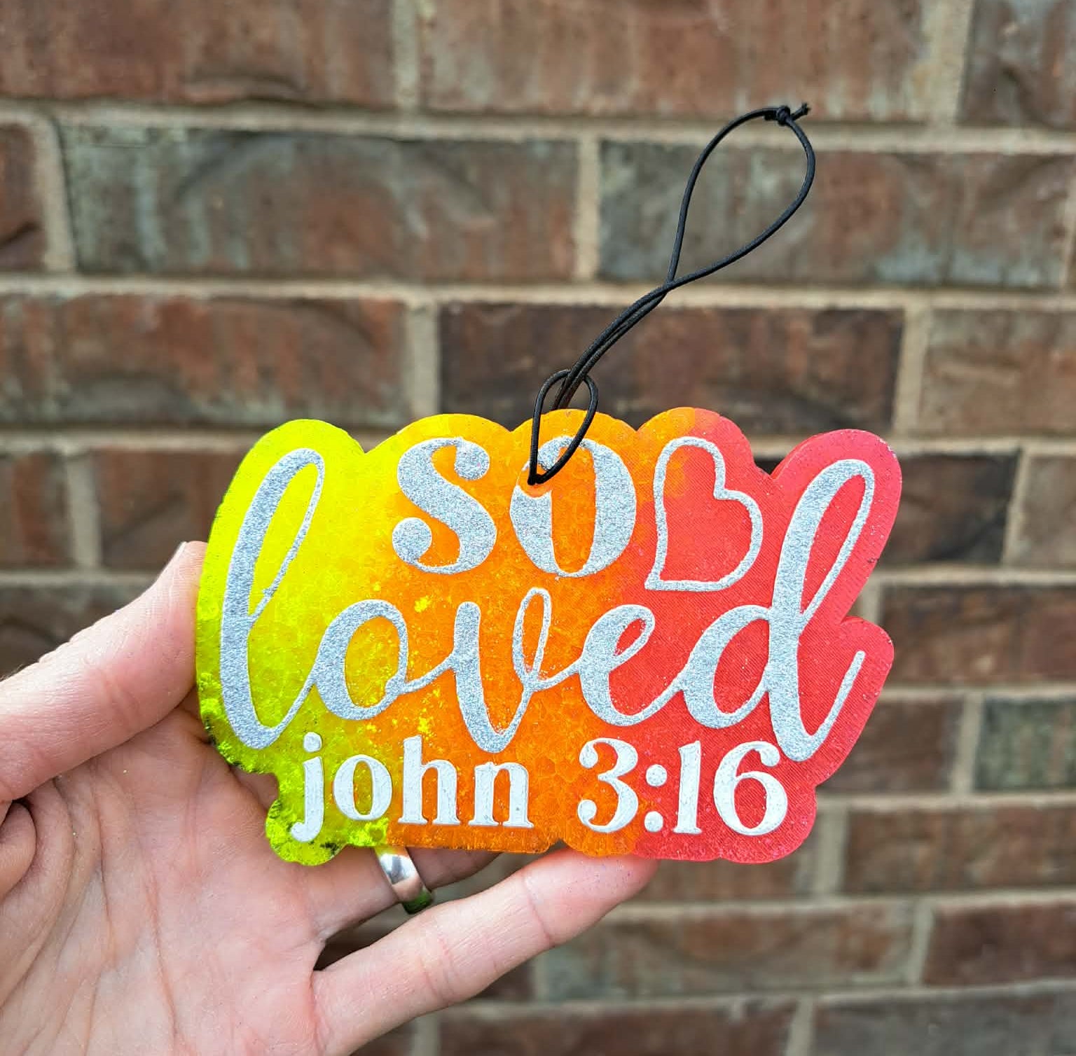 Colorful freshie with 'I am so loved' and 'John 3:16' text held against a brick wall.