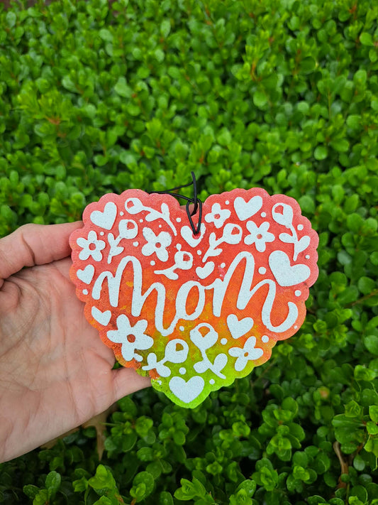 Heart-shaped freshie with 'mom' text held in front of green foliage