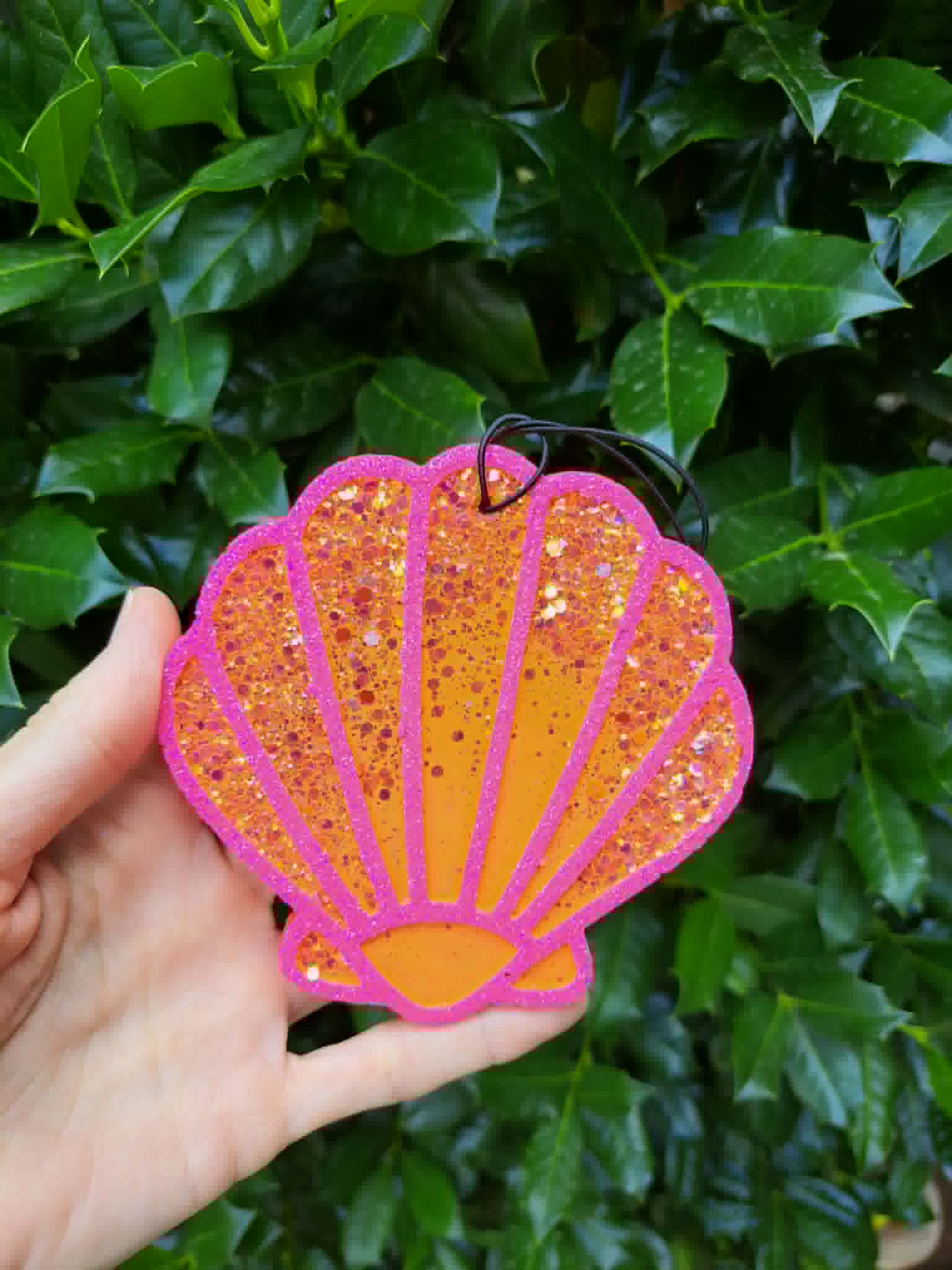 Hand holding a glittery pink and orange shell-shaped freshie against a green leafy background