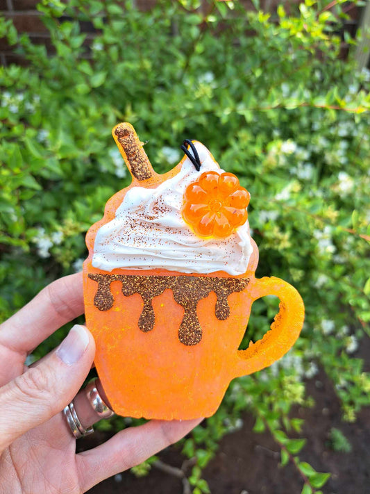 An orange air freshener mug with whipped cream and pumpkin decoration being held by a hand, with plants in the background.