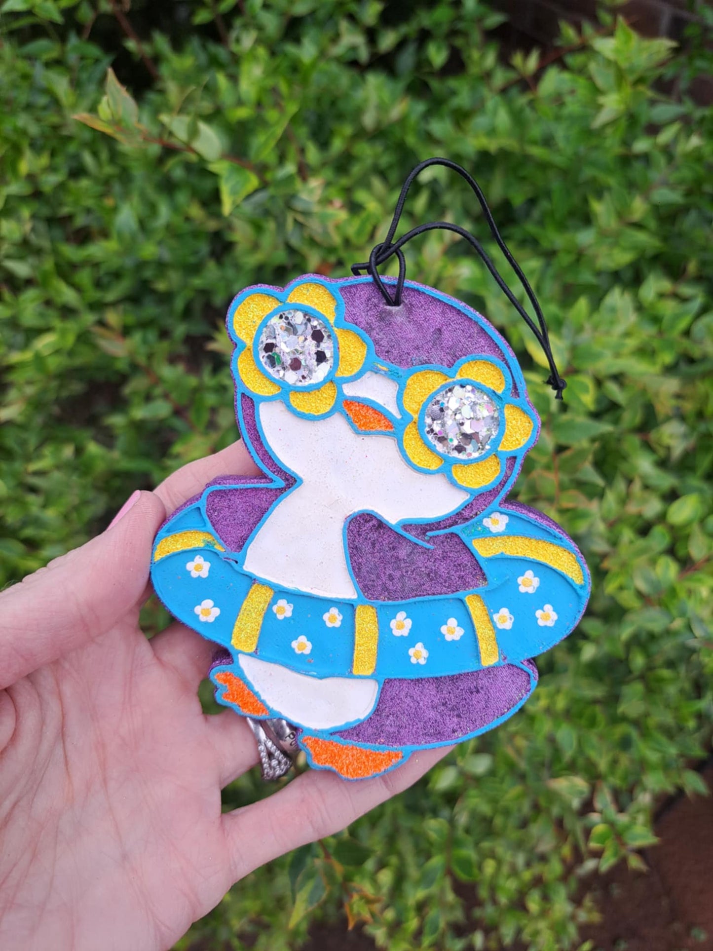 A handmade, painted air freshener in the shape of a floating penguin with blue and yellow colors and a black elastic string.