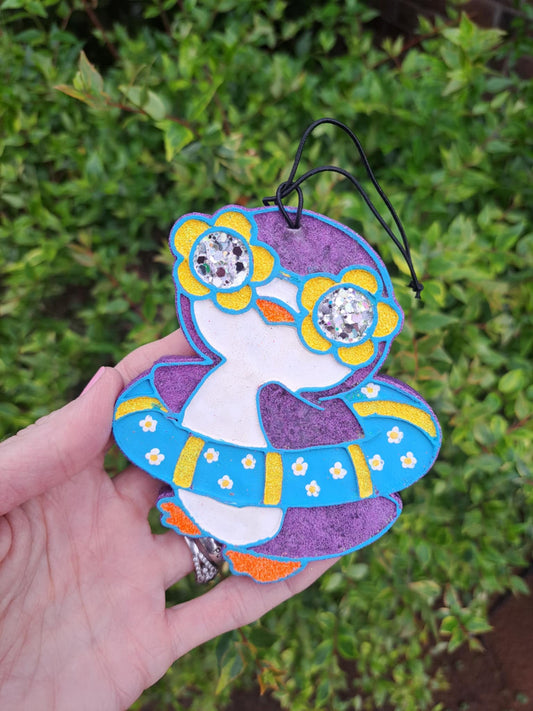 A handmade, painted air freshener in the shape of a floating penguin with blue and yellow colors and a black elastic string.