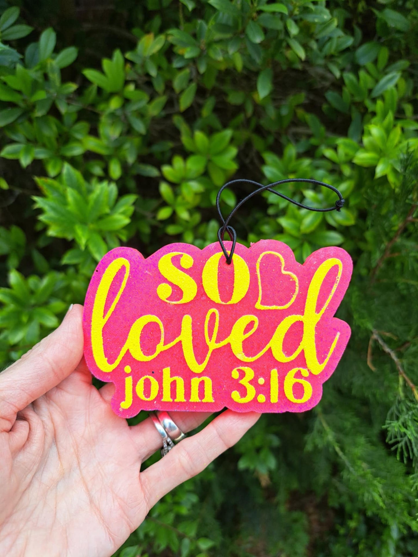 Pink and yellow 'So Loved' freshie with 'John 3:16' held in a hand against a green leafy background