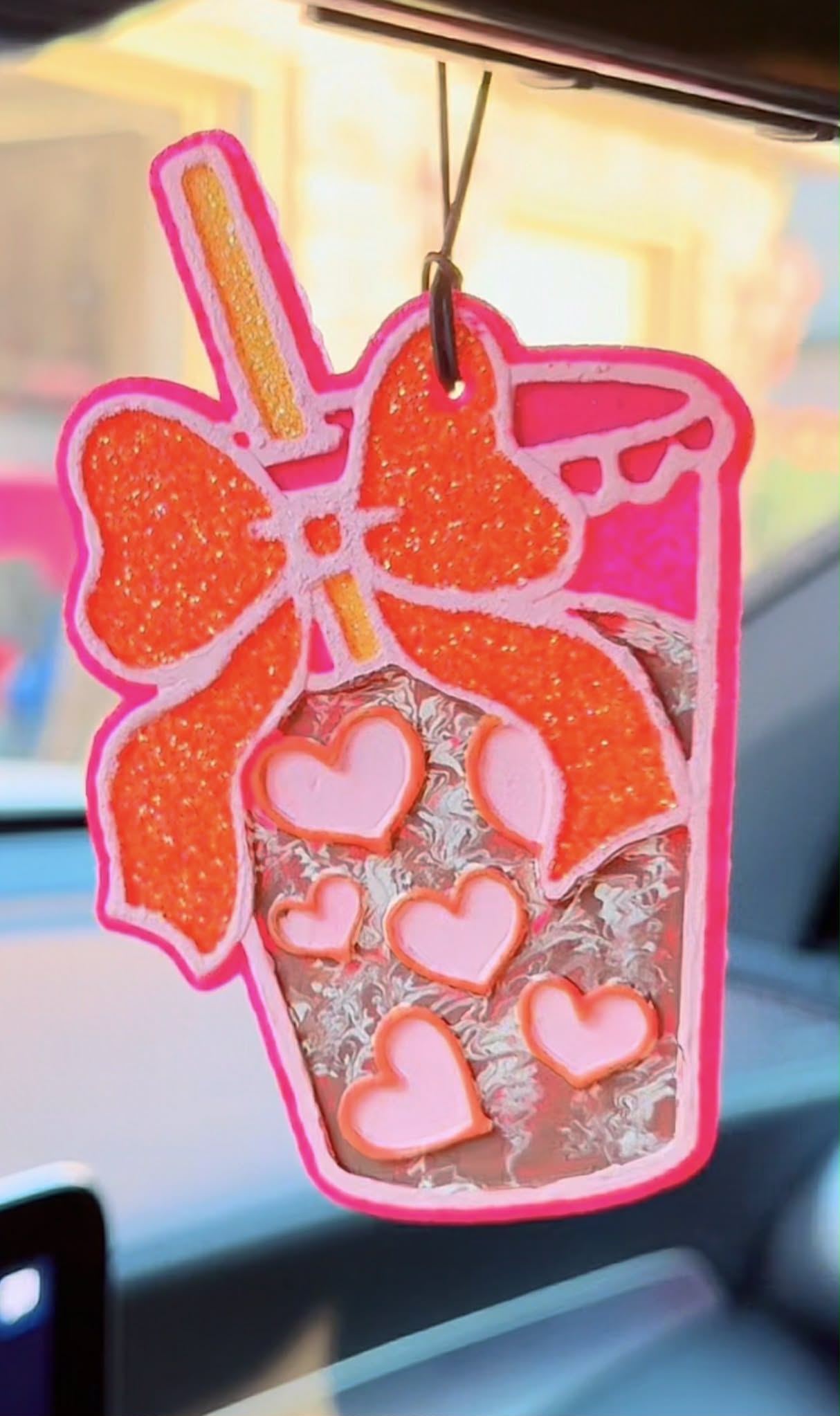 Car air freshener with an orange bow and heart designs hanging from a car's rearview mirror.