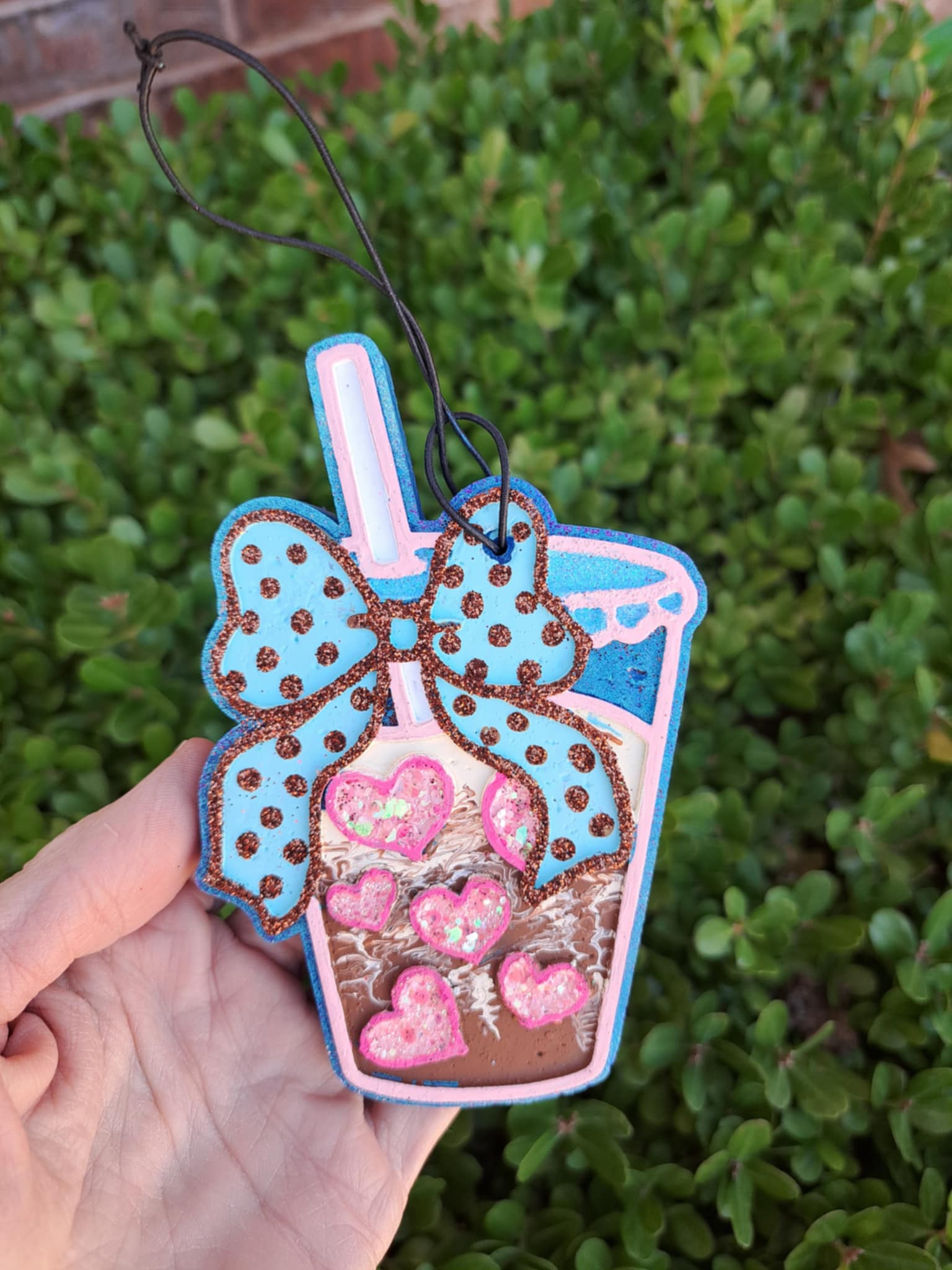 Decorative freshie shaped like a milkshake with a bow held in front of green foliage.