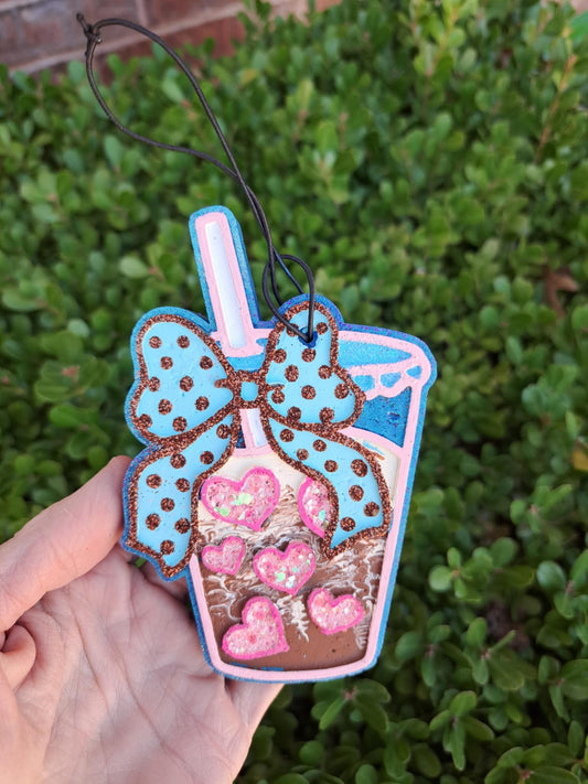 Decorative freshie shaped like a milkshake with a bow held in front of green foliage.