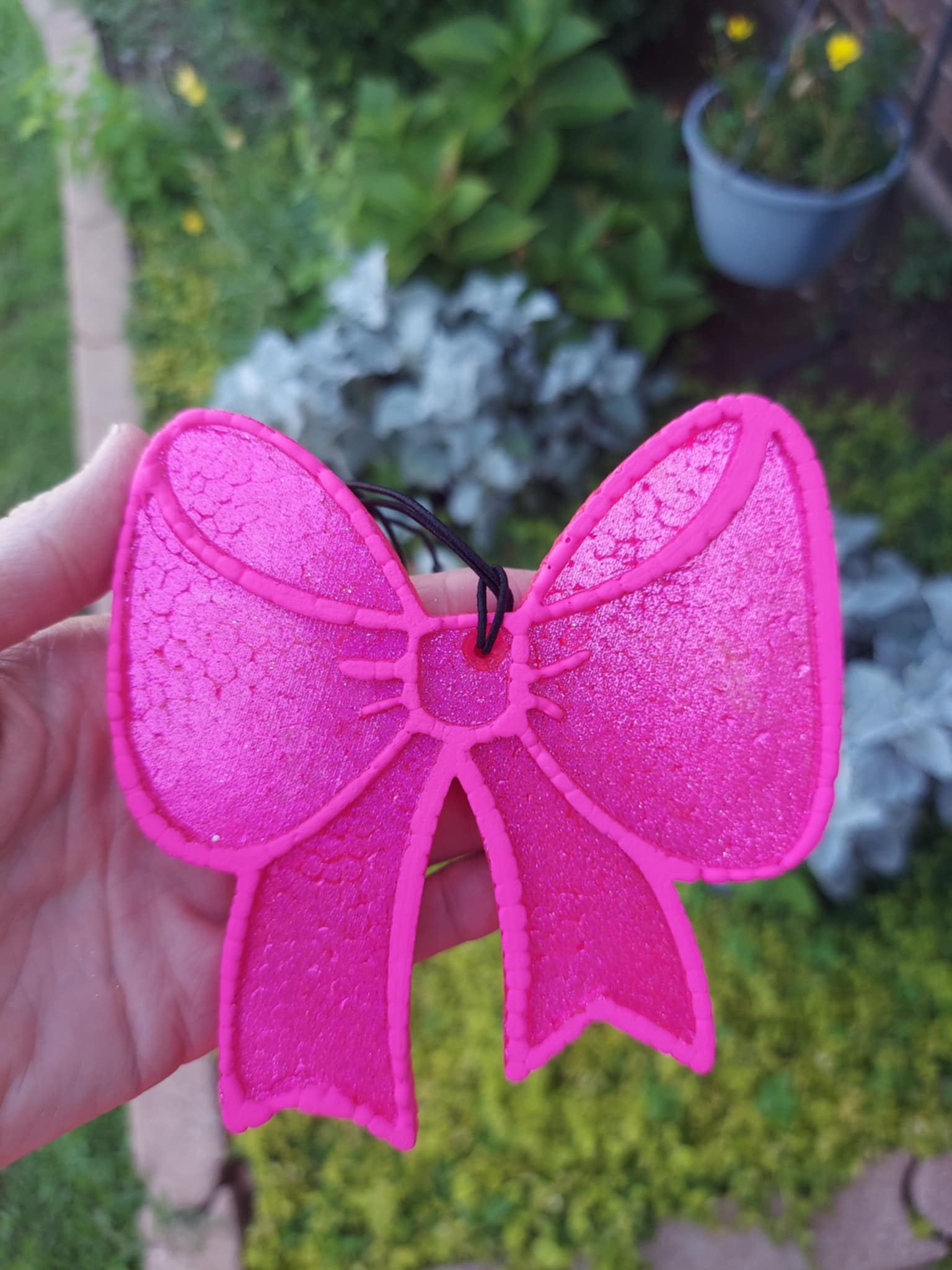 pink bow freshie held in a hand in front of flower garden
