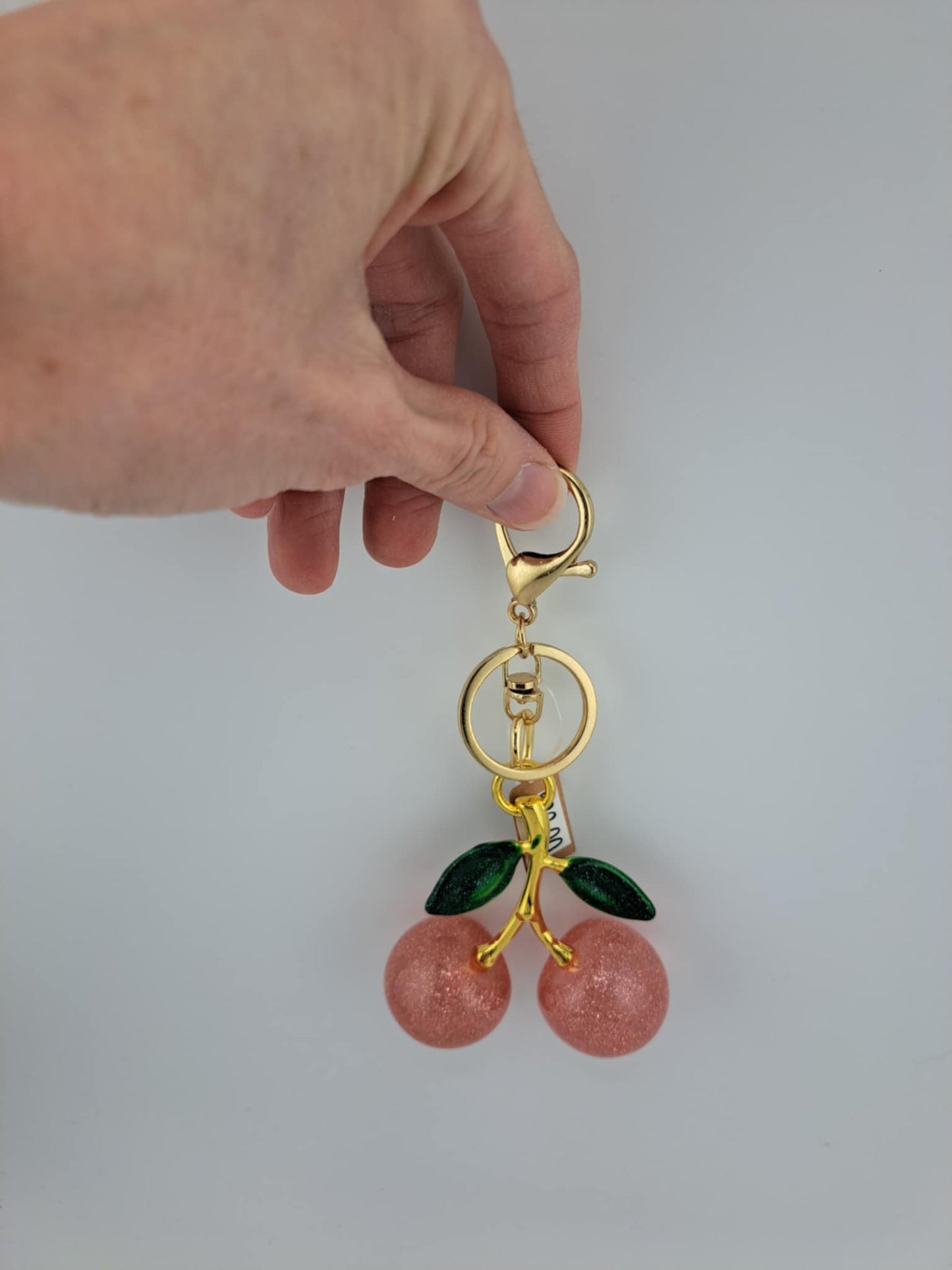 Gold keychain with cherry-shaped charms held by a hand against a light gray background