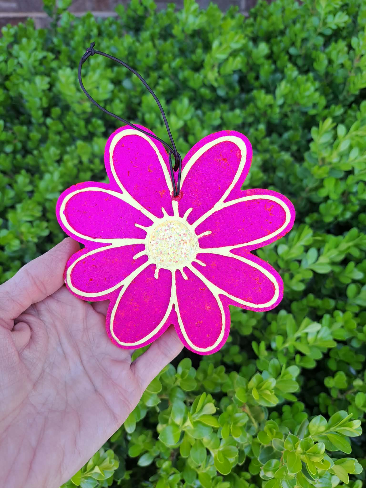 Hand holding a pink flower-shaped freshie with a green bush in the background