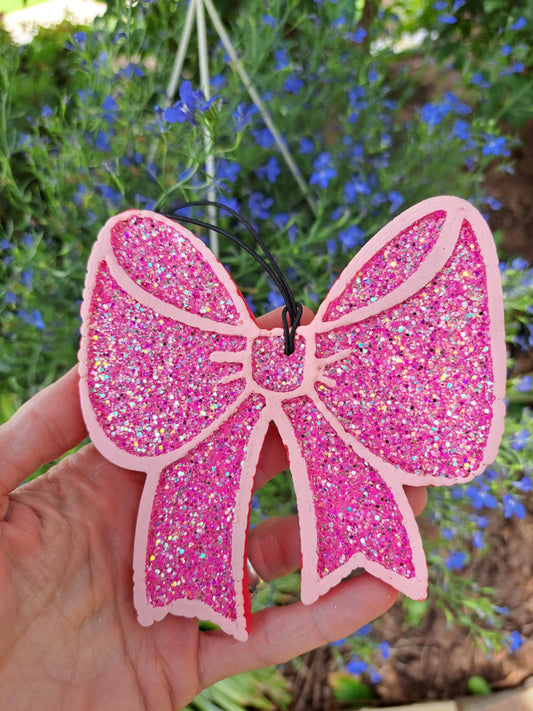 Pink glittery bow-shaped freshie held in front of a blurred natural background with blue flowers.