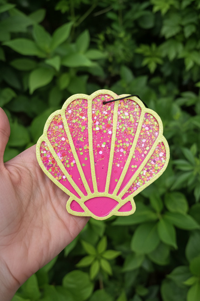 Pink and yellow glittery shell-shaped car air freshener held by a hand against a dark background