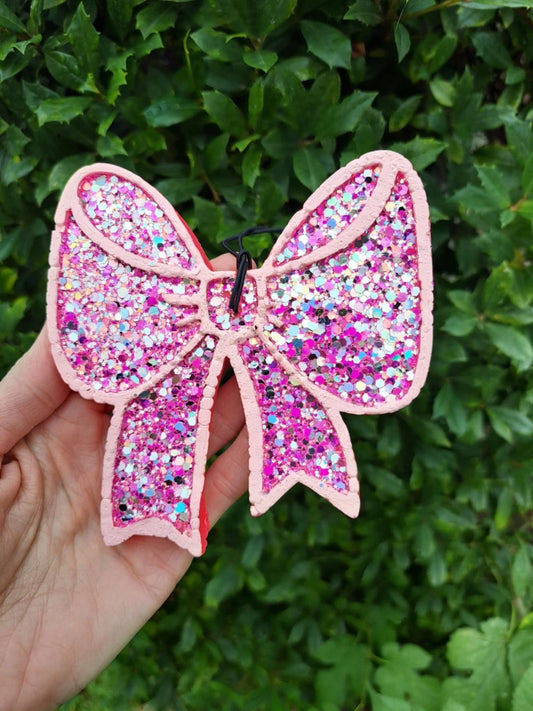 A hand holds a pink glitter bow-shaped freshie against a background of green leaves. The item is covered in pink, purple, and iridescent glitter with a light pink stitched border.