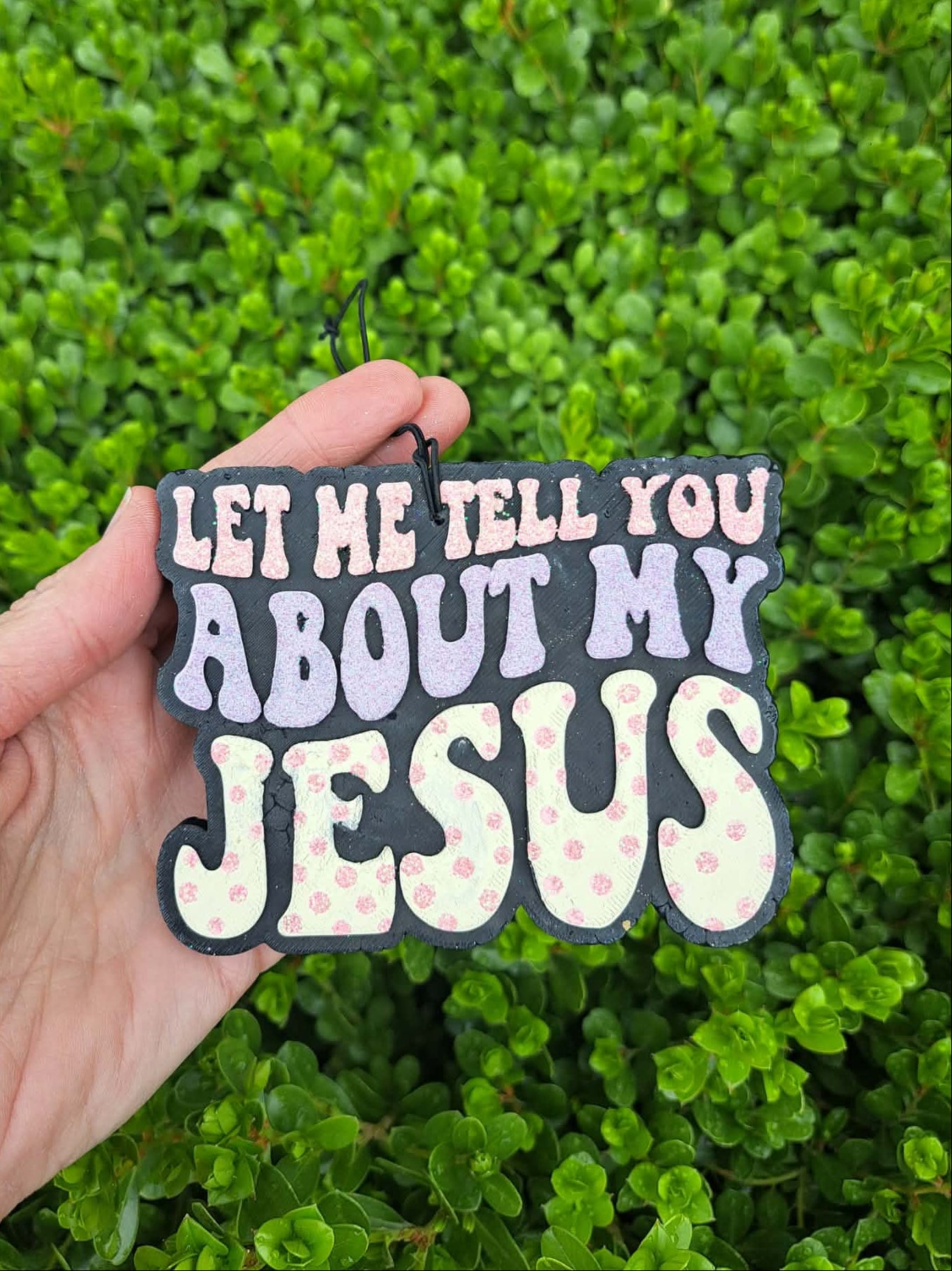 Hand holding a freshie with text 'Let me tell you about my Jesus' against a green leafy background