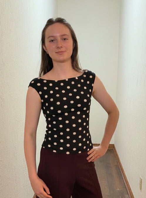 A woman stands in a narrow indoor hallway wearing a black sleeveless top with beige polka dots and dark burgundy wide-leg pants.