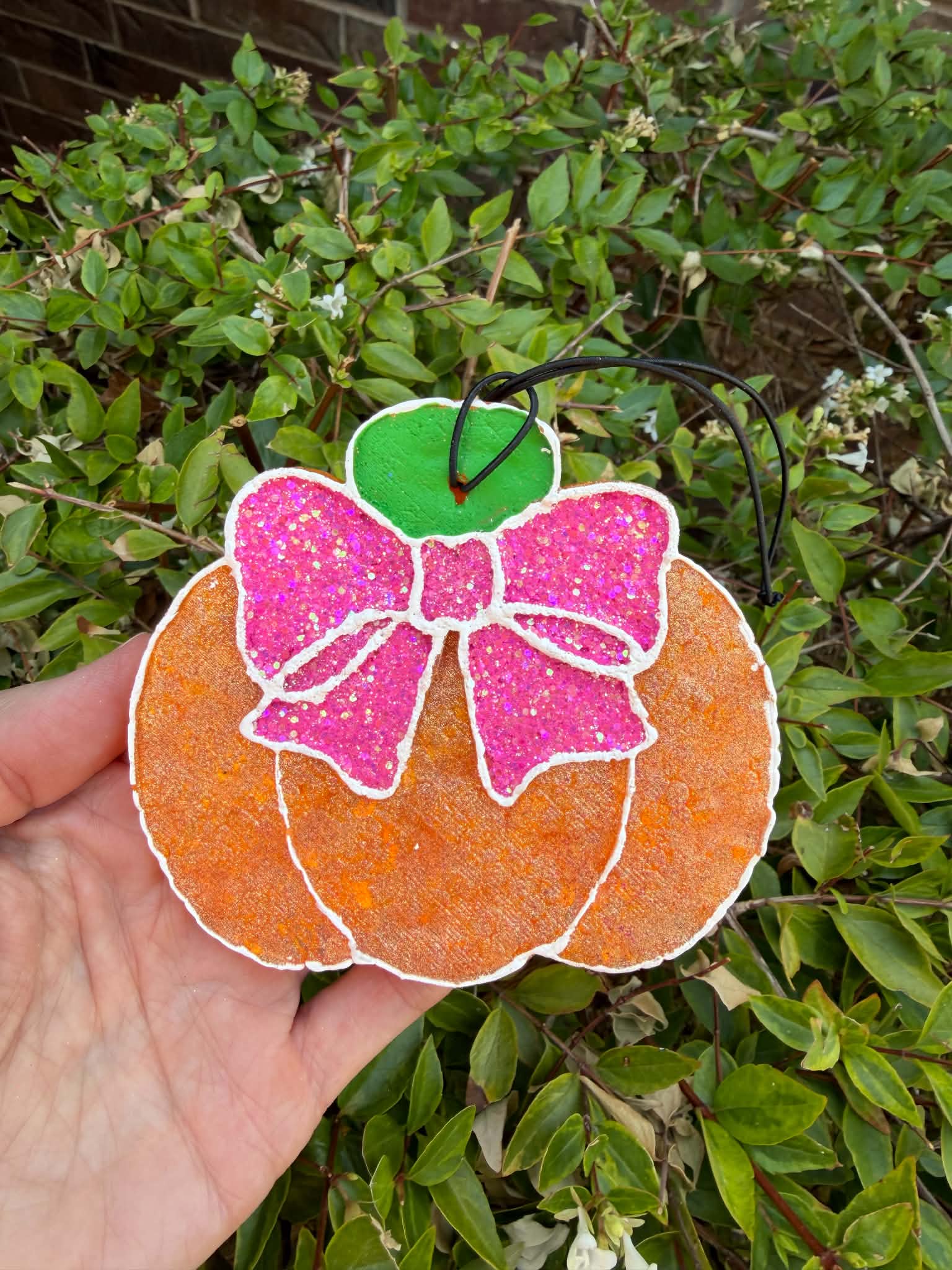 A hand holds a glittery pumpkin with a bright pink bow, green stem, and orange body against a background of green garden foliage.
