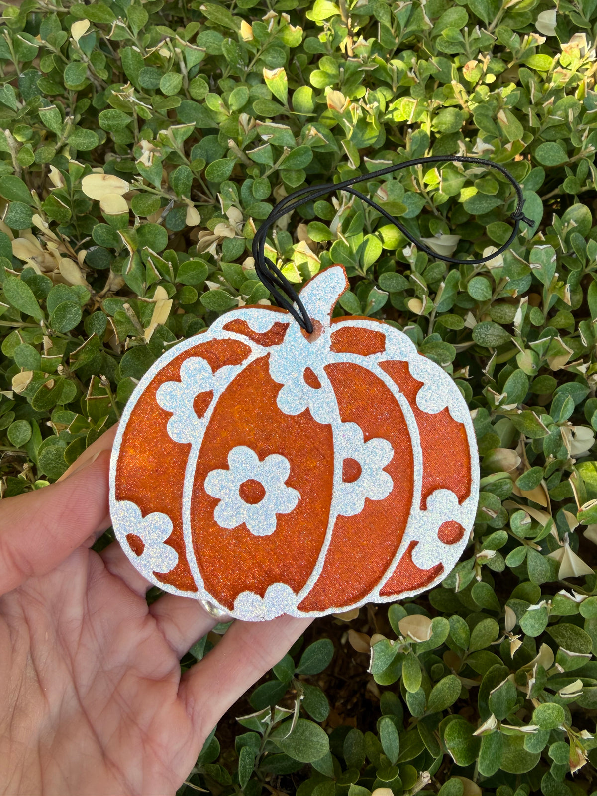 A hand holds a glittery orange pumpkin-shaped freshie with white floral patterns and a black hanging string, set against a background of green leafy plants.