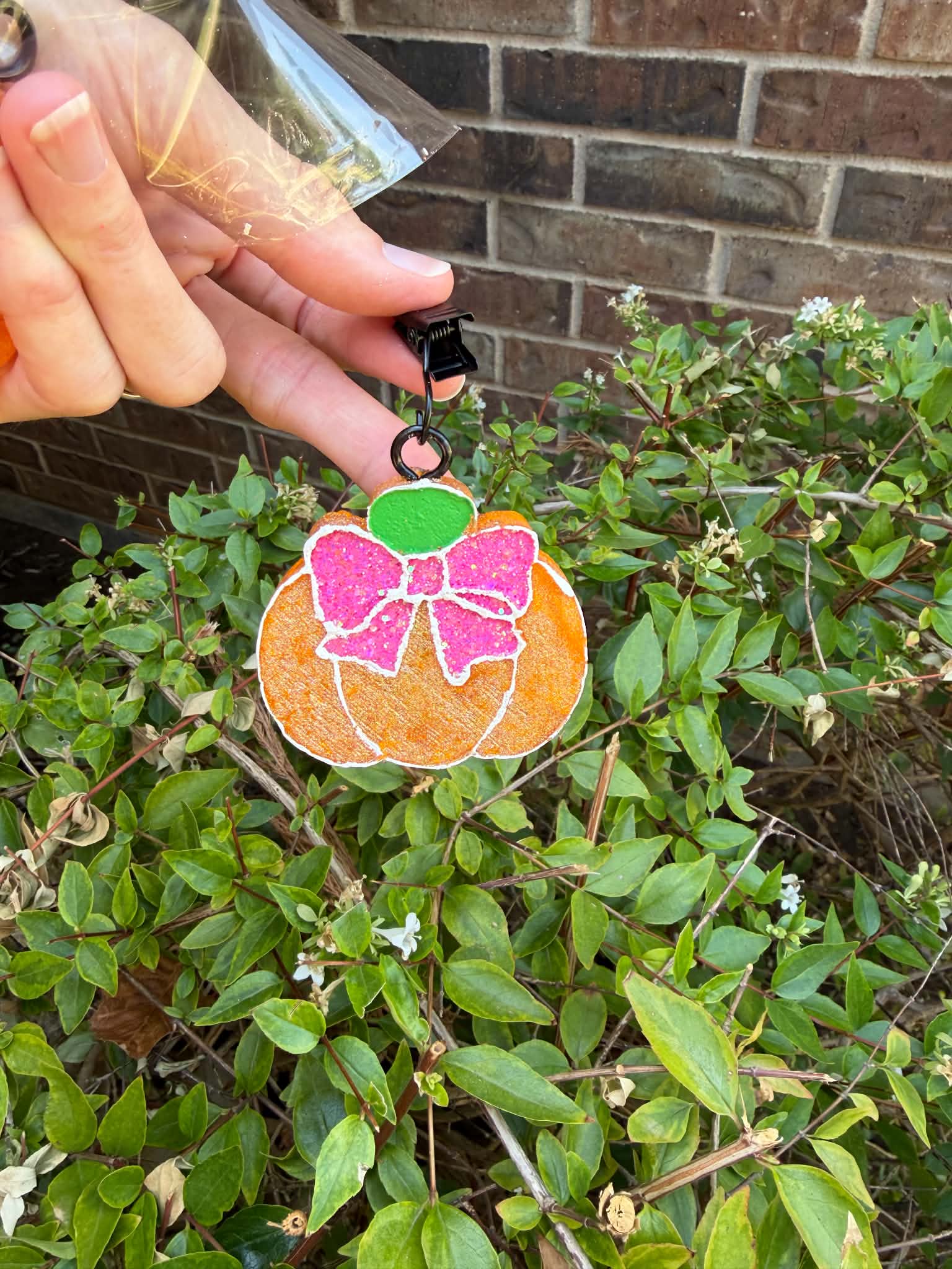 A hand holds a small orange pumpkin-shaped vent clip with a pink bow and green top in front of green bushes and a brick wall.