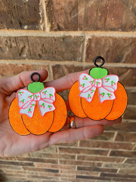 Two vent clips with a pumpkin and cherry print design bow, held in a hand against a brick wall background.