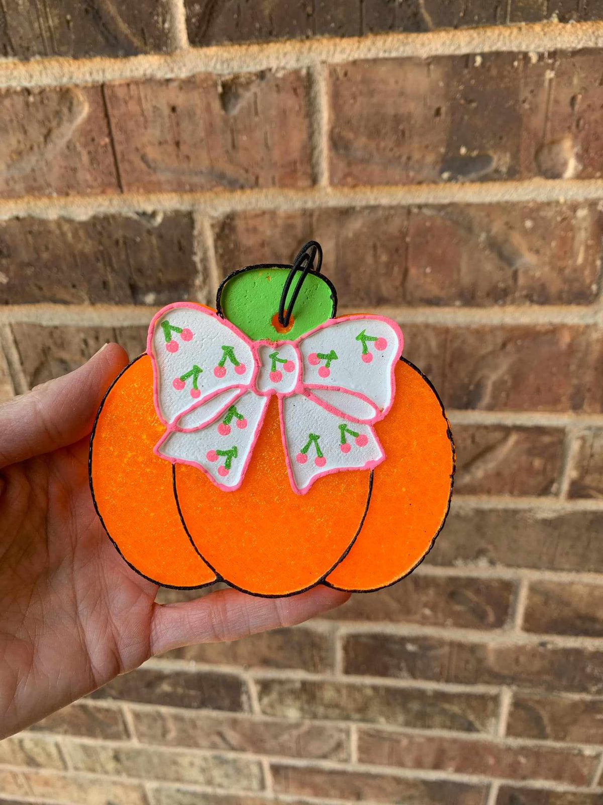 A hand holding a pumpkin-shaped air freshener with a cherry bow design against a brick wall background.