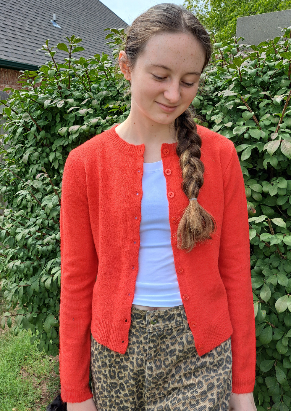 Person wearing a red cardigan over a white shirt with leopard print pants, standing in front of green bushes.