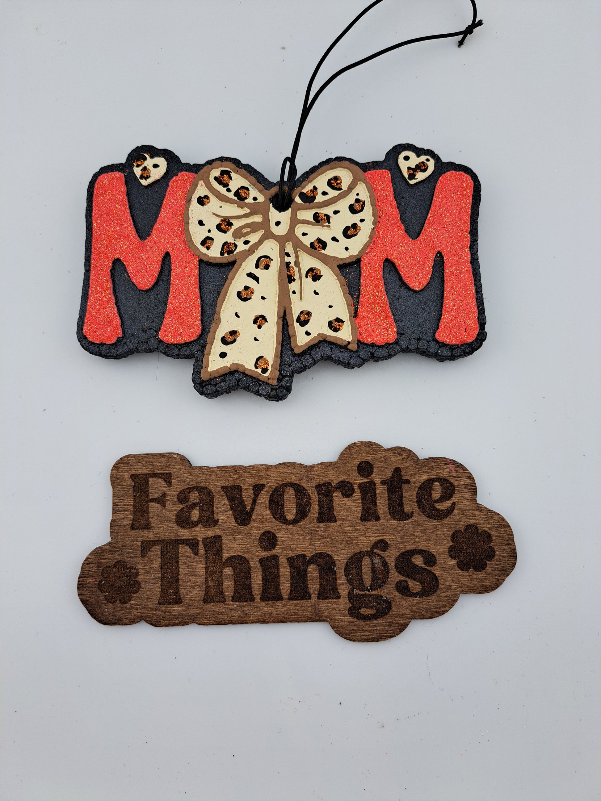 A handmade and painted air freshener with 'MOM' written in bold, leopard print design on a red background, with a bow. Below it, the phrase 'Favorite Things' is written in a simple font.