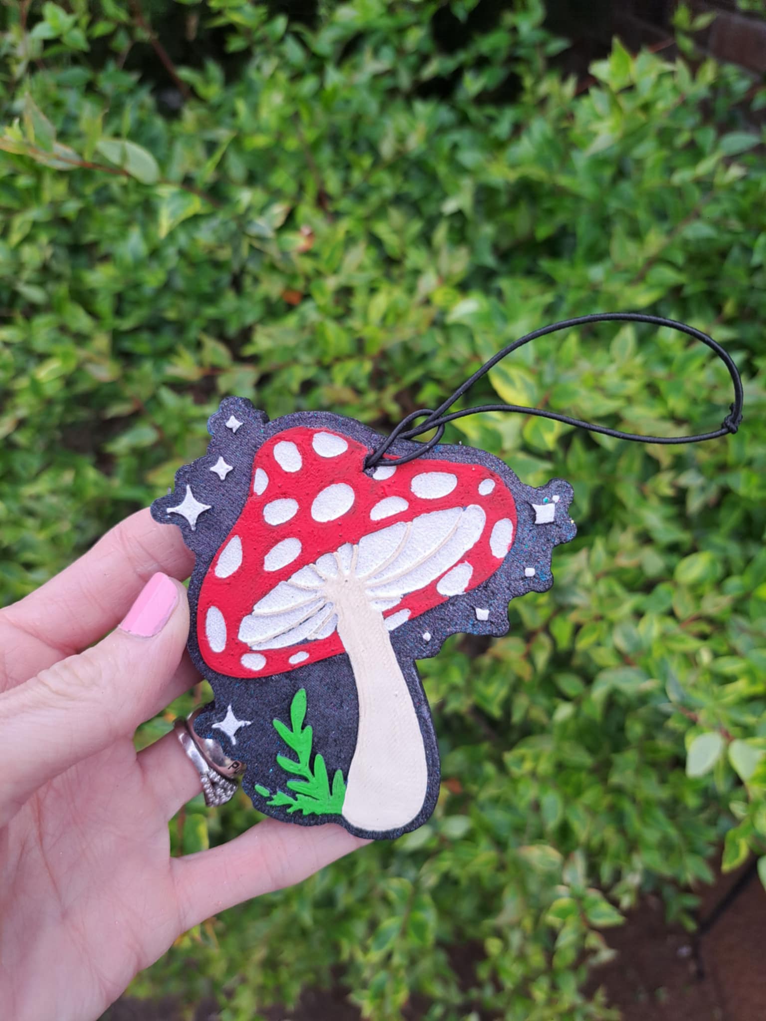 Hand holding a red and black handcrafted air freshener shaped like a mushroom with a toadstool design, featuring white spots and a white stem, attached to an elastic string.
