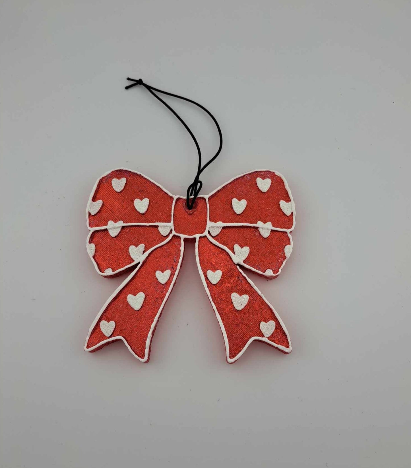 A red bow-shaped air freshener with white hearts pattern, hanging on black elastic string.