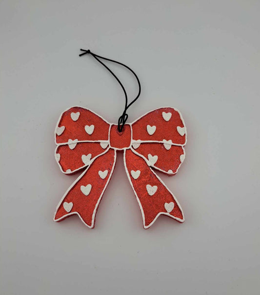 A red bow-shaped air freshener with white hearts pattern, hanging on black elastic string.