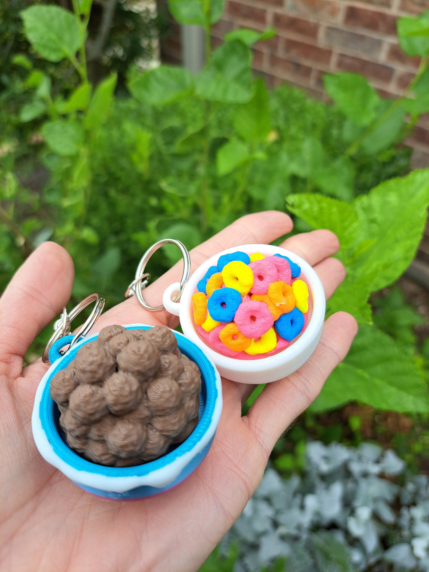 Hand holding two 3D printed cereal keychains against a green outdoor background
