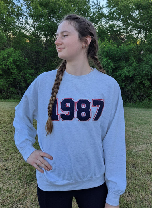 Person wearing a light blue sweatshirt with '1987' printed on it, standing outdoors with trees in the background.